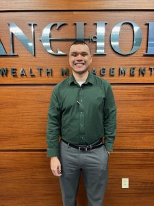 Young man in black shirt with grey pants standing in front of a paneld wall with the word Anchor Wealth Management in the background with an anchor through the work anchor